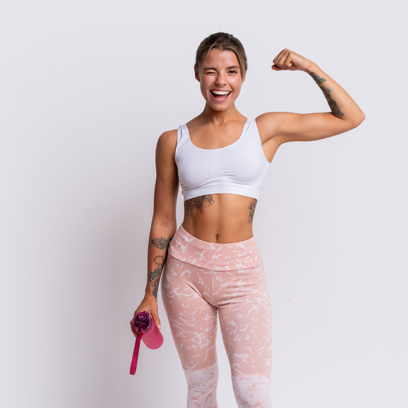 Fit blond woman with perfect smile in stylish sportswear looking at camera and holding bottle of water over white background. Demonstrate muscles.
